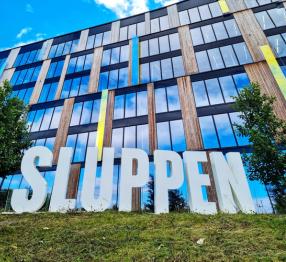 Big letters spelling out "SLUPPEN", on a field of green grass in front of a tall building made with a lot of glass