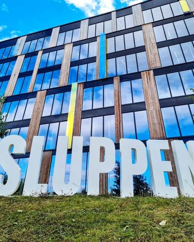 Big letters spelling out "SLUPPEN", on a field of green grass in front of a tall building made with a lot of glass