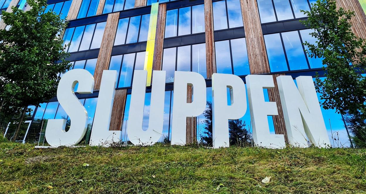 Big letters spelling out "SLUPPEN", on a field of green grass in front of a tall building made with a lot of glass