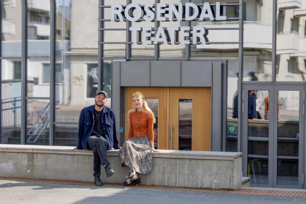 A man and a woman sitting on a bench outside of a new and fresh theatre building. The front wall of the theatre is made of glass