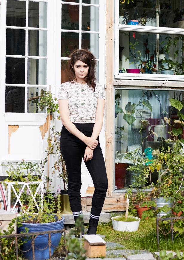 a young woman standing outside a house, with a variety of plants visible through the windows, suggesting a green, vibrant, and cozy atmosphere inside. She stands near a small garden or porch area, surrounded by potted plants, and appears thoughtful or introspective as she gazes at the camera.