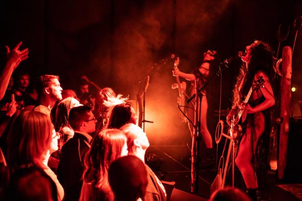 A female black-metal band performing on stage. The lighting in the room is red, and there's fog. The crowd loves it.