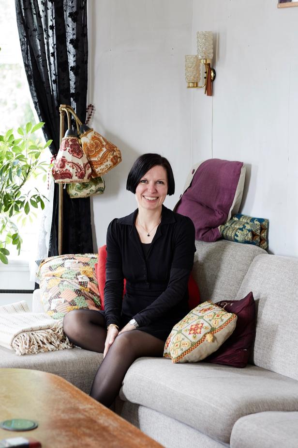 A woman sitting comfortably on a sofa in a cozy living room. She is smiling and wearing a black outfit, along with dark tights and soft shoes. Behind her are several colorful pillows and bags hanging on the wall. The setting has a warm, inviting atmosphere with plants and soft lighting, suggesting a homey and relaxed environment.