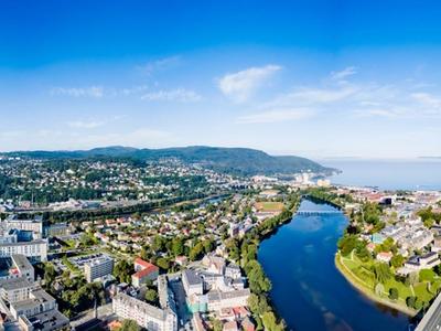 A panoramic aerial view of Trondheim, Norway, on a clear and sunny day. The Nidelva river winds through the city, reflecting the blue sky. In the foreground, urban buildings and green parks are visible, with the NTNU Gløshaugen campus and St. Olavs Hospital standing out. The city extends towards rolling hills covered with houses and forests. To the right, the Trondheimsfjord stretches into the horizon, with a distant ship visible on the calm waters. The sky is bright blue with a few wispy clouds.