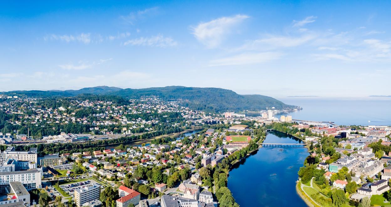A panoramic aerial view of Trondheim, Norway, on a clear and sunny day. The Nidelva river winds through the city, reflecting the blue sky. In the foreground, urban buildings and green parks are visible, with the NTNU Gløshaugen campus and St. Olavs Hospital standing out. The city extends towards rolling hills covered with houses and forests. To the right, the Trondheimsfjord stretches into the horizon, with a distant ship visible on the calm waters. The sky is bright blue with a few wispy clouds.