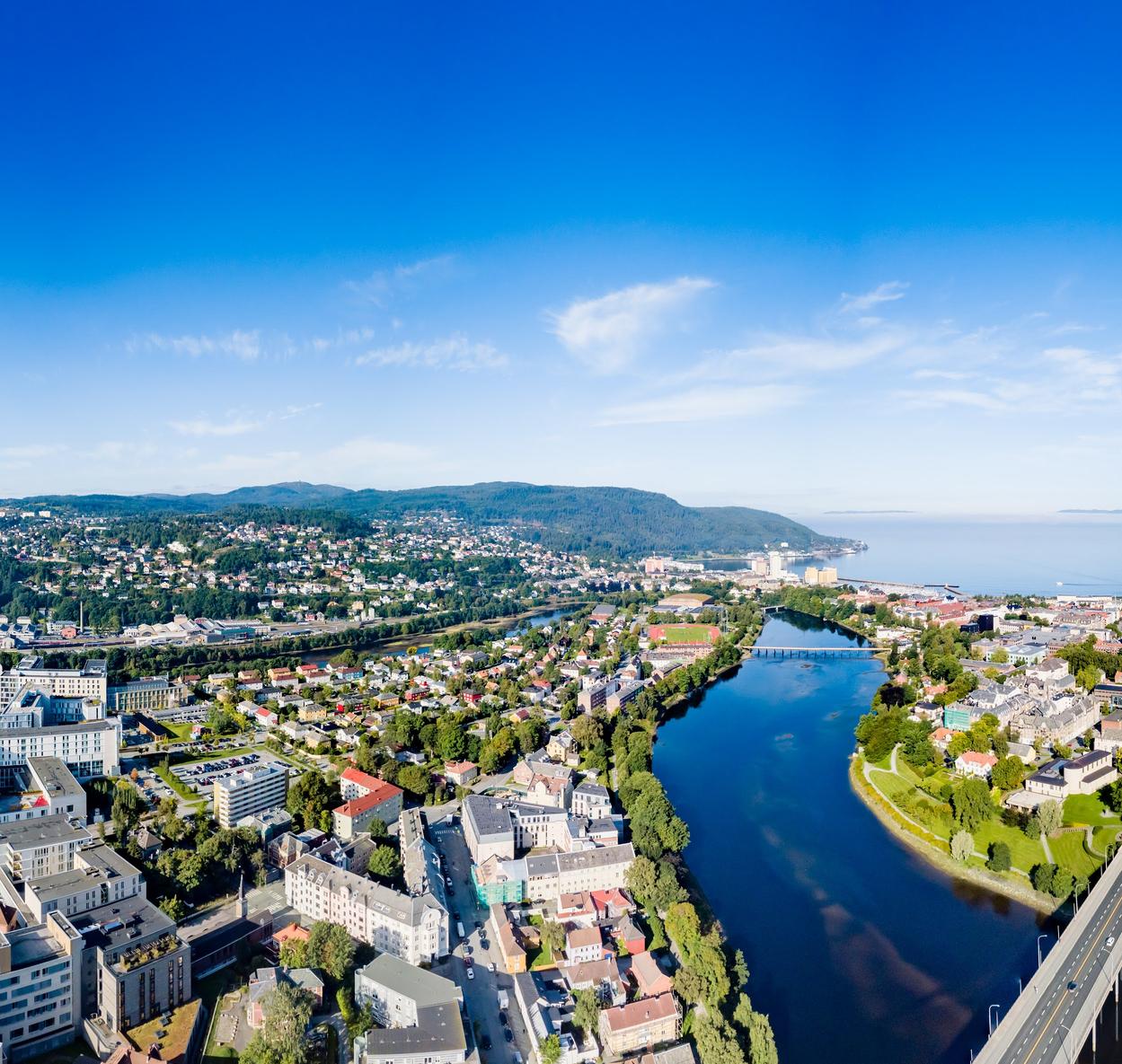 A panoramic aerial view of Trondheim, Norway, on a clear and sunny day. The Nidelva river winds through the city, reflecting the blue sky. In the foreground, urban buildings and green parks are visible, with the NTNU Gløshaugen campus and St. Olavs Hospital standing out. The city extends towards rolling hills covered with houses and forests. To the right, the Trondheimsfjord stretches into the horizon, with a distant ship visible on the calm waters. The sky is bright blue with a few wispy clouds.