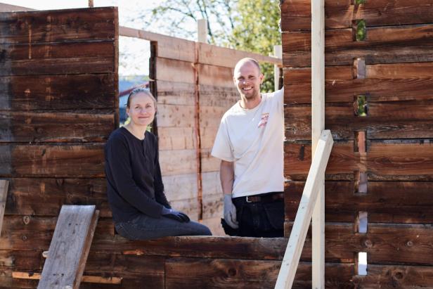 two people, a man and a woman, sitting in or near a wooden structure under construction. They are both smiling and seem to be taking a break or enjoying the moment while working on the building. The wooden walls around them are partially constructed, and there’s some daylight coming through, suggesting that it’s a sunny day.