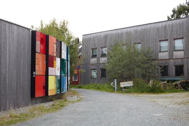 modern building with a wall adorned with a colorful display, possibly made up of various doors or panels in vibrant colors. The path leads toward the building, and it seems to have a natural, green environment around it.