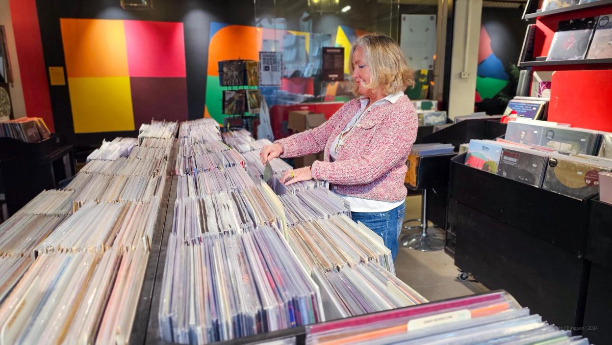 The same woman in a pink tweed jacket smiling as she looks at records. The store's interior is filled with rows of vinyls, music posters, and bright wall art.