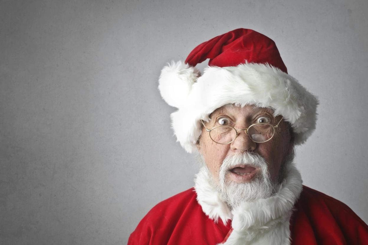 An elderly man with a Santa Claus outfit and a surprised expression, wearing a Santa hat and round glasses.