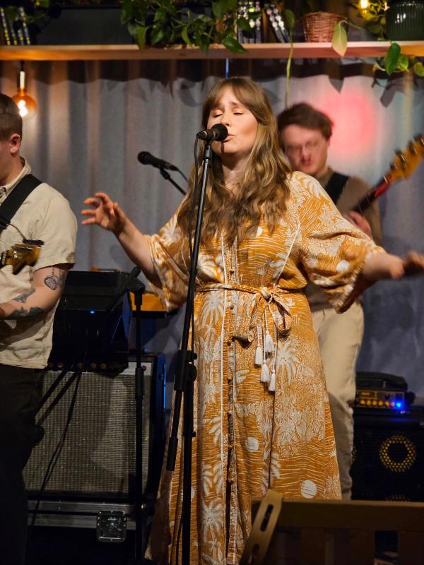 A female singer with long, wavy blonde hair performs on stage, wearing a flowing mustard-yellow dress with white floral patterns. She sings into a microphone with her eyes closed and expressive hand gestures. Behind her, a guitarist in a beige outfit plays, partially blurred. The stage has warm lighting, with potted plants and a cozy ambiance in the background, suggesting an intimate live music venue.