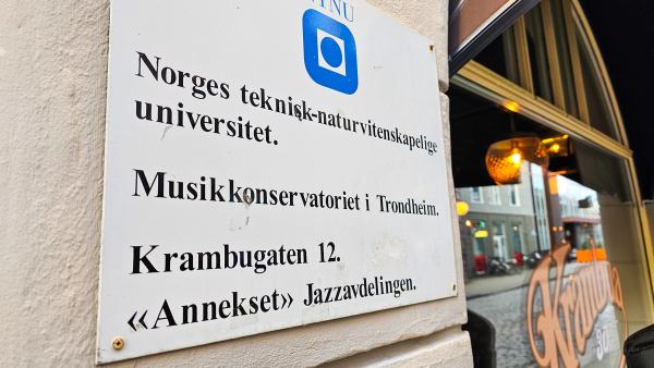 A close-up of a white sign on a wall, displaying the text "Norges teknisk-naturvitenskapelige universitet. Musikkonservatoriet i Trondheim. Krambugaten 12. ‘Anneks’ Jazzavdelingen." The sign has the NTNU logo at the top, and a café window reflecting lights is visible in the background.