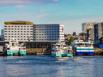 Several boats docked at a harbor in Trondheim, with a modern building in the background. The boats, identified by their distinct colors and markings, are part of the local transportation network, likely used for ferry services. The photo has a slight blur effect on the background, focusing on the boats in the foreground.