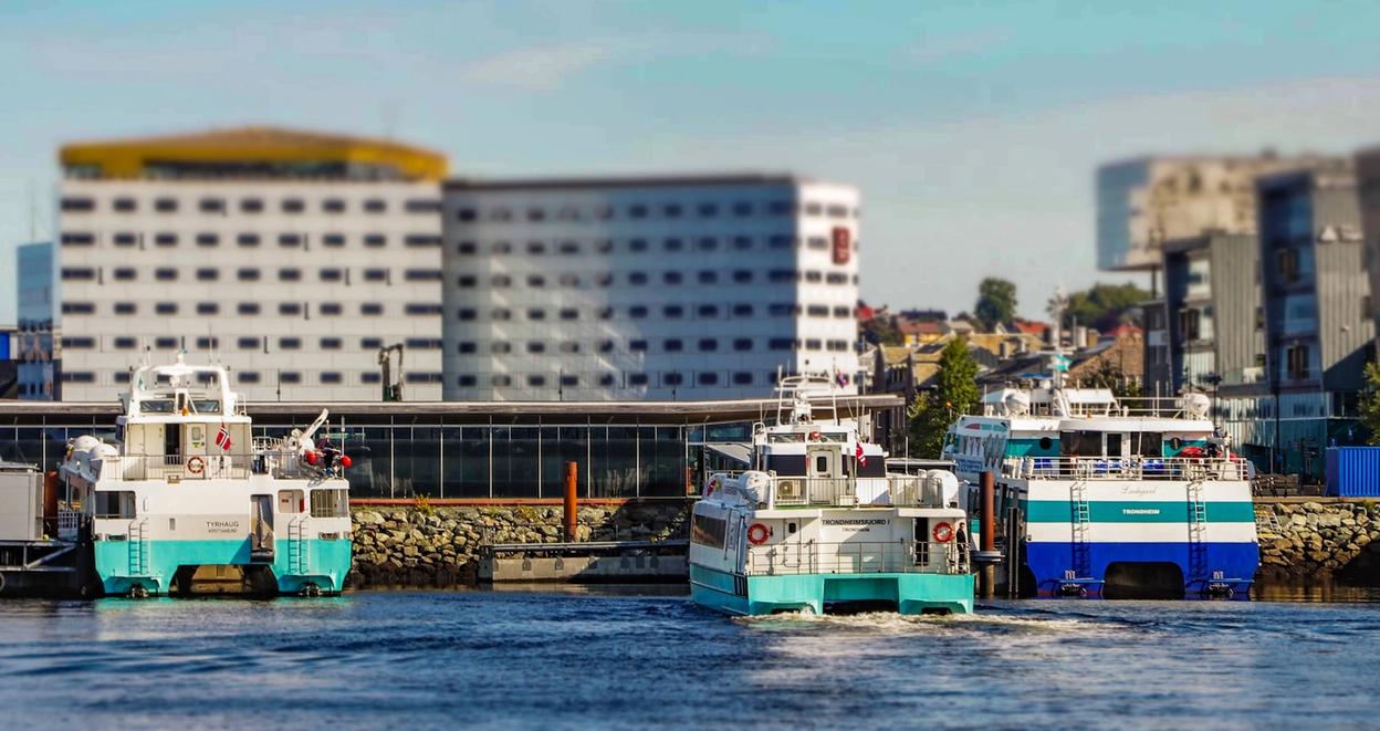 Several boats docked at a harbor in Trondheim, with a modern building in the background. The boats, identified by their distinct colors and markings, are part of the local transportation network, likely used for ferry services. The photo has a slight blur effect on the background, focusing on the boats in the foreground.
