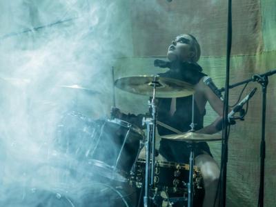 A drummer performing on stage surrounded by smoke, with intense focus and energy. The drummer is dressed in dark clothing and has a dramatic pose, playing a drum kit under dim lighting, adding to the atmospheric, electrifying vibe of the performance.*