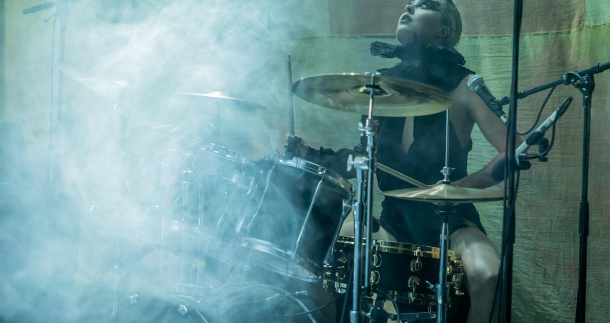 A drummer performing on stage surrounded by smoke, with intense focus and energy. The drummer is dressed in dark clothing and has a dramatic pose, playing a drum kit under dim lighting, adding to the atmospheric, electrifying vibe of the performance.*