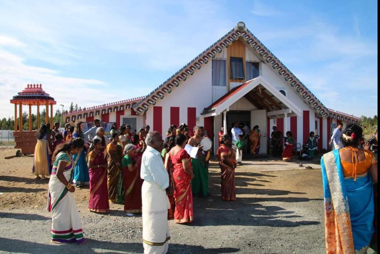 This image shows a vibrant group of people in traditional Indian attire, gathered outside a building with distinctive red and white stripes. The setting appears to be a cultural or religious event, with individuals dressed in colorful sarees, standing in front of a temple or community centre.