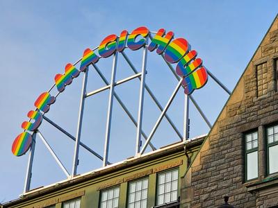 A vibrant rainbow-colored sign reading 'OH MAGIC HOUSE' mounted on a metal frame above a historic stone building with a clock on its facade in Trondheim. The sky in the background is clear blue.