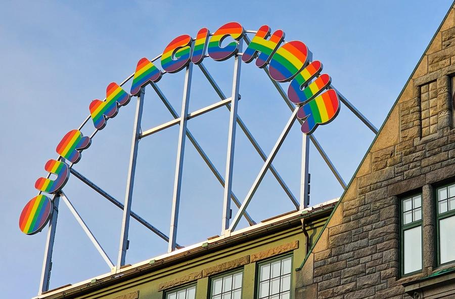 A vibrant rainbow-colored sign reading 'OH MAGIC HOUSE' mounted on a metal frame above a historic stone building with a clock on its facade in Trondheim. The sky in the background is clear blue.