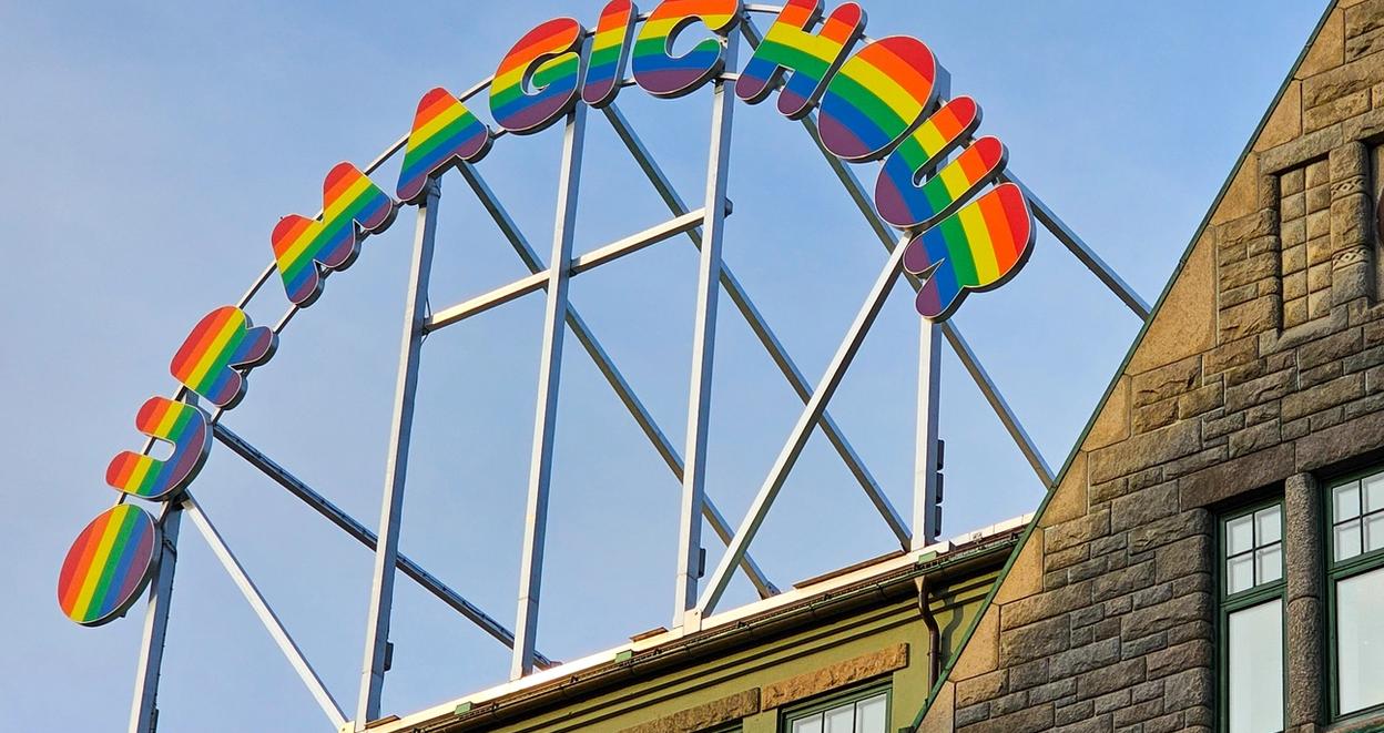 A vibrant rainbow-colored sign reading 'OH MAGIC HOUSE' mounted on a metal frame above a historic stone building with a clock on its facade in Trondheim. The sky in the background is clear blue.