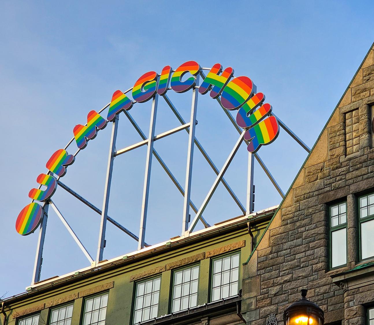 A vibrant rainbow-colored sign reading 'OH MAGIC HOUSE' mounted on a metal frame above a historic stone building with a clock on its facade in Trondheim. The sky in the background is clear blue.