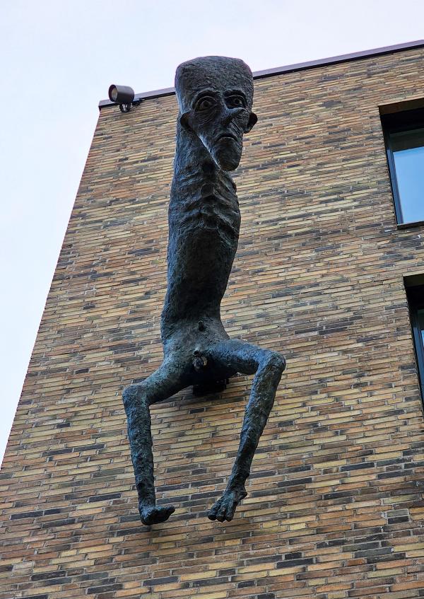 Close-up of K-U-K Sculpture. A close-up of a bronze sculpture featuring a tall, gaunt figure with an elongated head and thin limbs protruding from the side of a brick building in Trondheim.