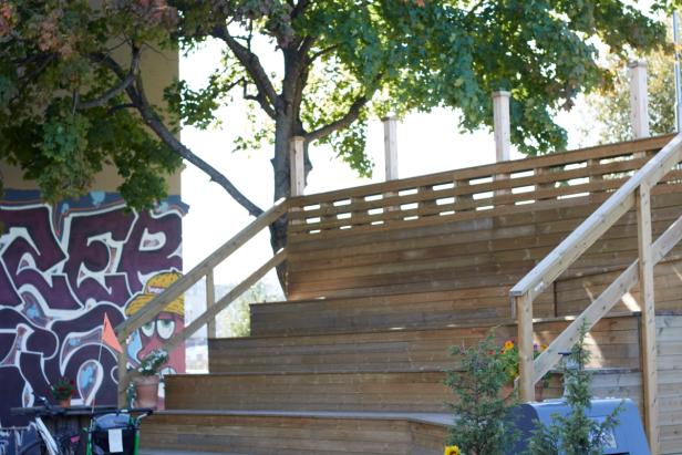 a set of wooden stairs or seating steps, leading up to a platform, surrounded by trees. In the background, there is graffiti artwork on the wall, which adds a colorful contrast to the natural and wooden elements in the scene. The space appears to be an outdoor public or communal area, likely designed for gatherings or relaxing. The plants and the warm lighting from the sun suggest a pleasant environment.