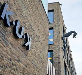 K-U-K Art Center Building. A brick building with large black letters spelling 'K-U-K' on its facade. A surreal sculpture of a long-limbed creature extends from the wall. The sky is overcast.