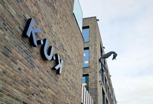 K-U-K Art Center Building. A brick building with large black letters spelling 'K-U-K' on its facade. A surreal sculpture of a long-limbed creature extends from the wall. The sky is overcast.