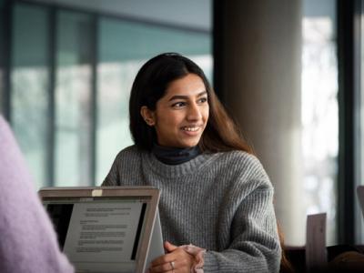 A young woman in a grey sweater smiling while engaged in a conversation, sitting at a table with a laptop in a modern indoor setting.