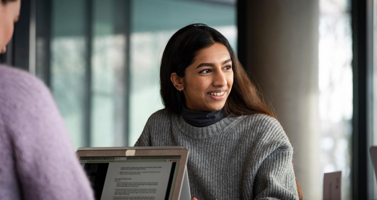 A young woman in a grey sweater smiling while engaged in a conversation, sitting at a table with a laptop in a modern indoor setting.