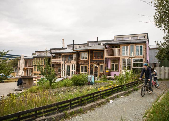 A row of colorful, eco-friendly houses in Svartlamon, Trondheim, built with repurposed materials and featuring large windows and wooden balconies. A gravel path runs alongside a small fenced-off green area with wild plants. A person wearing a blue jacket and helmet rides a bicycle with a child following behind, adding to the vibrant, community-focused atmosphere.