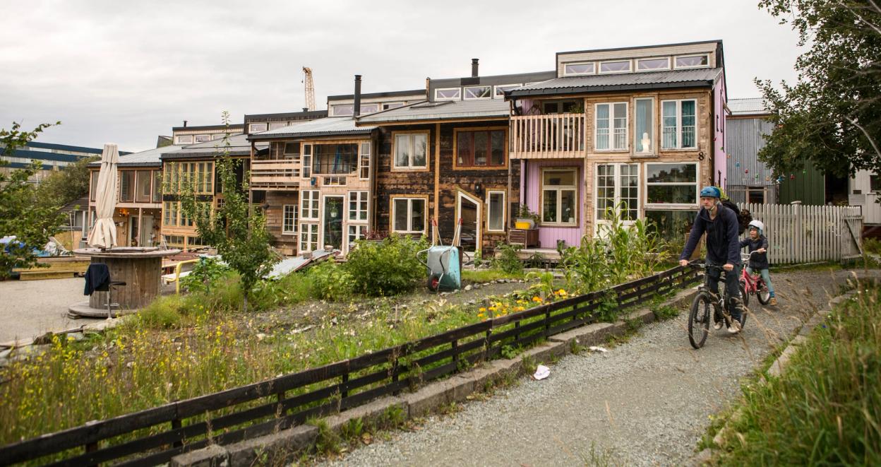 A row of colorful, eco-friendly houses in Svartlamon, Trondheim, built with repurposed materials and featuring large windows and wooden balconies. A gravel path runs alongside a small fenced-off green area with wild plants. A person wearing a blue jacket and helmet rides a bicycle with a child following behind, adding to the vibrant, community-focused atmosphere.