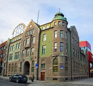 A historic green and stone building with a tower, arched windows, and green sculptures of animals climbing its façade. In the background, a red modern theatre hall contrasts with the older architecture.