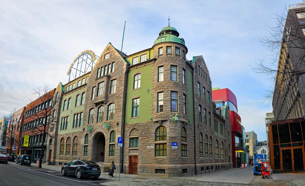 A historic green and stone building with a tower, arched windows, and green sculptures of animals climbing its façade. In the background, a red modern theatre hall contrasts with the older architecture.