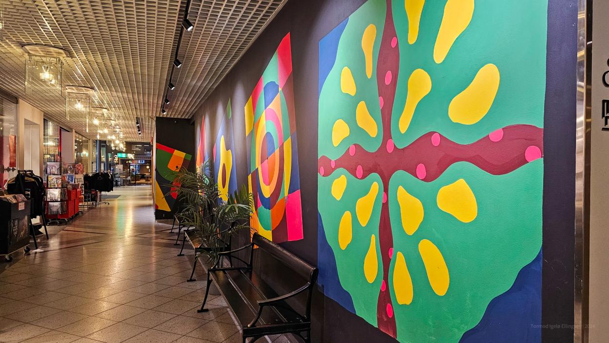 A long corridor with large, colorful murals on a black wall. The geometric and abstract artwork is vibrant, and there are benches and potted plants along the hallway.