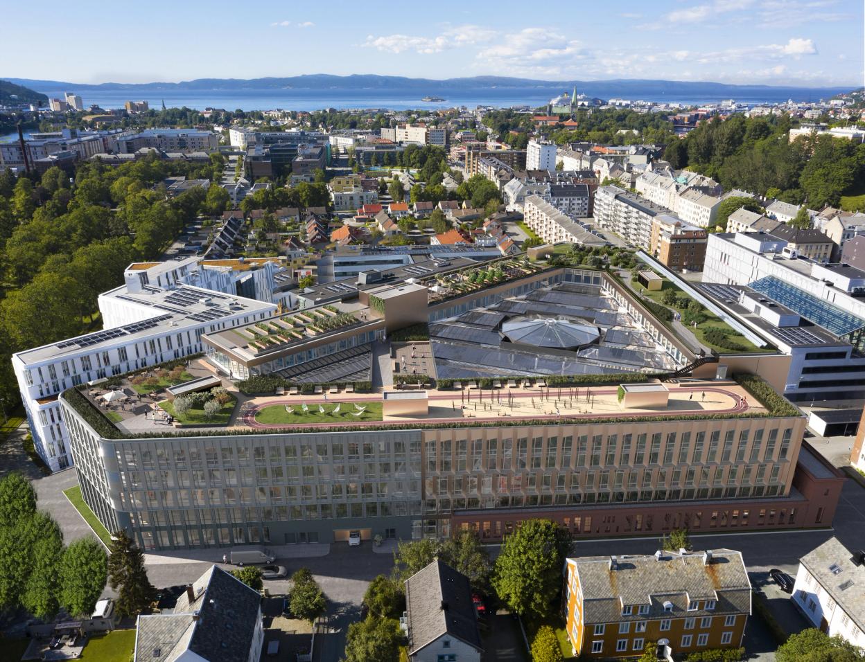 Teknostallen in Trondheim, Norway. The modern building features a green rooftop with garden spaces, solar panels, and an outdoor track. Its unique pentagonal design includes an open central courtyard with a glass roof. Surrounding the building is a mix of residential and commercial structures, with lush greenery and tree-lined streets. In the background, the Trondheimsfjord and Nidaros Cathedral are visible under a clear blue sky.