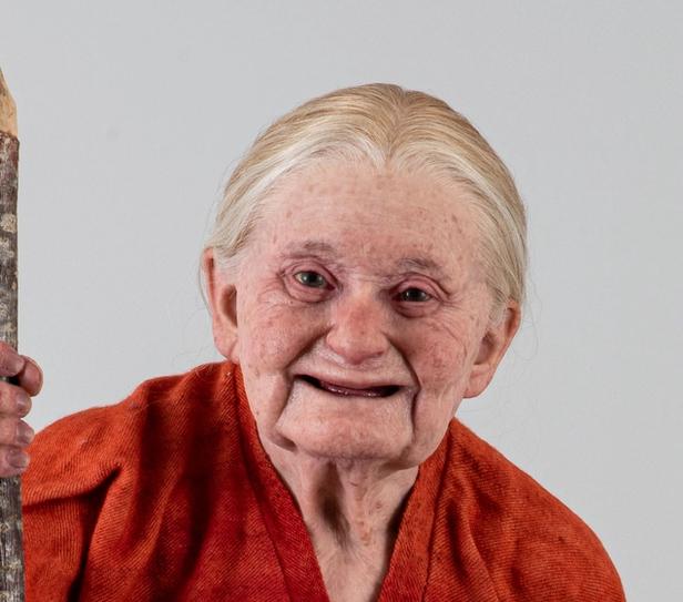 An elderly woman wearing a traditional, bright red long dress stands confidently with a walking stick in hand. Her silver hair is tied back, and her warm expression reflects wisdom and strength. She is barefoot, adding a natural and grounded element to the image. The simplicity of her attire and the serene background highlight her presence and character.