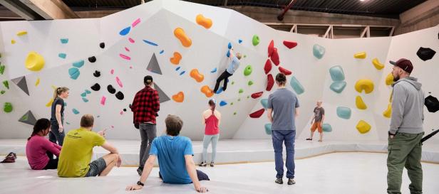 A group of people standing in front of a bouldering course. One person is already on the bouldering course, making his way towards the top.