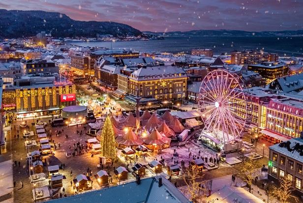 Aerial view of a Christmas market in a snowy city square at dusk, illuminated by festive lights. A large Ferris wheel with pink and purple lights stands prominently, surrounded by wooden market stalls, glowing tents, and a tall Christmas tree wrapped in golden lights. Snowflakes are gently falling, adding to the winter atmosphere. The background features a harbor, mountains, and a colorful sunset sky, enhancing the scenic beauty of the location.