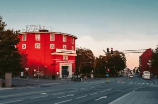 A round, red building, labeled "Studentersamfundet," stands prominently against a soft evening sky. The building has a distinctive circular shape and is framed by trees and street lights. The street in front of the building is quiet, with traffic lights indicating a calm moment during the day. The architecture is unique, and the warm, golden light adds a nostalgic and inviting atmosphere to the scene. The photo suggests this is a landmark or community center in the area.