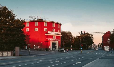 A round, red building, labeled "Studentersamfundet," stands prominently against a soft evening sky. The building has a distinctive circular shape and is framed by trees and street lights. The street in front of the building is quiet, with traffic lights indicating a calm moment during the day. The architecture is unique, and the warm, golden light adds a nostalgic and inviting atmosphere to the scene. The photo suggests this is a landmark or community center in the area.