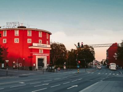 A round, red building, labeled "Studentersamfundet," stands prominently against a soft evening sky. The building has a distinctive circular shape and is framed by trees and street lights. The street in front of the building is quiet, with traffic lights indicating a calm moment during the day. The architecture is unique, and the warm, golden light adds a nostalgic and inviting atmosphere to the scene. The photo suggests this is a landmark or community center in the area.
