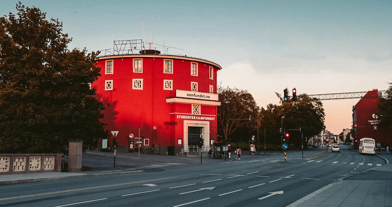 A round, red building, labeled "Studentersamfundet," stands prominently against a soft evening sky. The building has a distinctive circular shape and is framed by trees and street lights. The street in front of the building is quiet, with traffic lights indicating a calm moment during the day. The architecture is unique, and the warm, golden light adds a nostalgic and inviting atmosphere to the scene. The photo suggests this is a landmark or community center in the area.