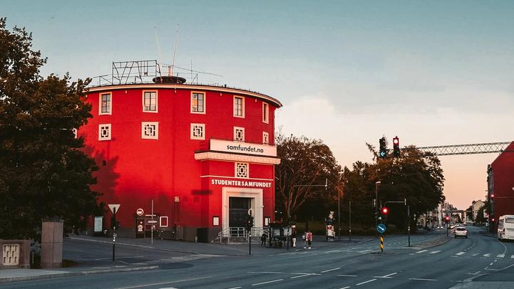 A round, red building, labeled "Studentersamfundet," stands prominently against a soft evening sky. The building has a distinctive circular shape and is framed by trees and street lights. The street in front of the building is quiet, with traffic lights indicating a calm moment during the day. The architecture is unique, and the warm, golden light adds a nostalgic and inviting atmosphere to the scene. The photo suggests this is a landmark or community center in the area.