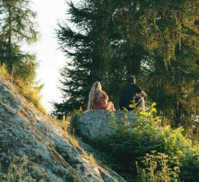 A couple sitting on a rock in a park on a hill. The weather is beautiful and the plants are green and lucious