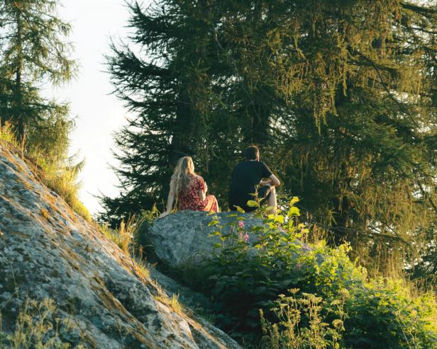 A couple sitting on a rock in a park on a hill. The weather is beautiful and the plants are green and lucious