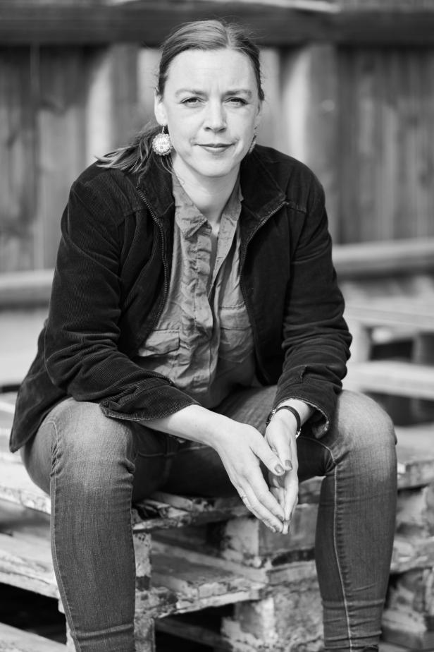 This black-and-white photo shows a woman sitting on a stack of wooden pallets. She is dressed casually in a button-up shirt and a jacket, with her hands placed on her knees. The background appears slightly blurred but suggests an outdoor setting. The woman's expression seems focused and calm, giving the photo a grounded and thoughtful feel.