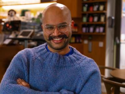 A man wearing a blue sweater, smiling inside of a coffee-shop. There are books in shelves behind him.