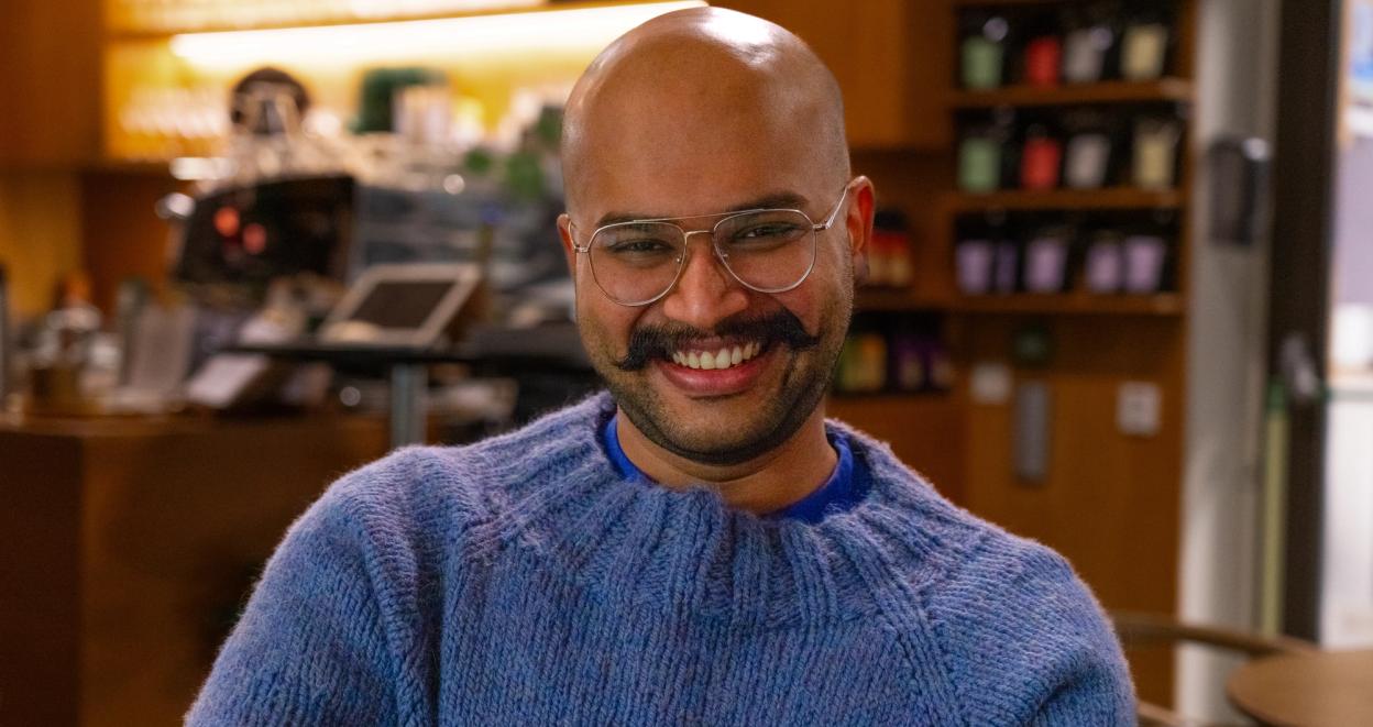 A man wearing a blue sweater, smiling inside of a coffee-shop. There are books in shelves behind him.